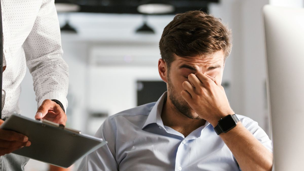 Homem cansado no escritório, com a mão no rosto, enquanto um colega mostra uma prancheta — sinal de esgotamento de fim de ano